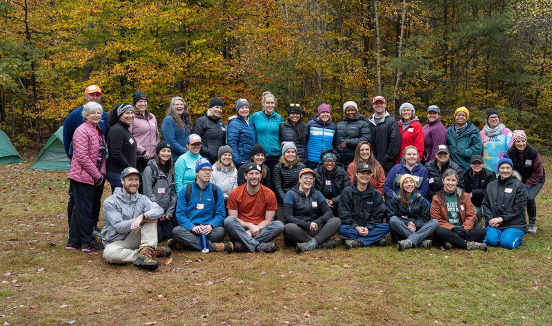 The image shows a large group of people posing for a photo outdoors. They are dressed in casual clothing, and many are wearing hats and jackets, suggesting it might be a cool day. The background features trees with colorful autumn foliage, indicating the photo was taken in the fall season.